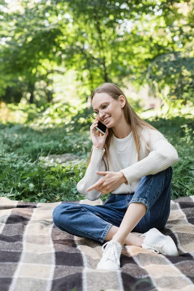 Fröhliche Frau mit Brille, die auf dem Smartphone spricht, während sie auf karierten Karos sitzt — Stockfoto