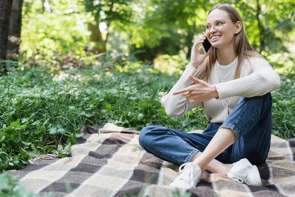 Joyful woman in glasses talking on smartphone while sitting on checkered plaid — Stock Photo