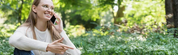 Joyful woman in glasses talking on smartphone in green park, banner - foto de stock