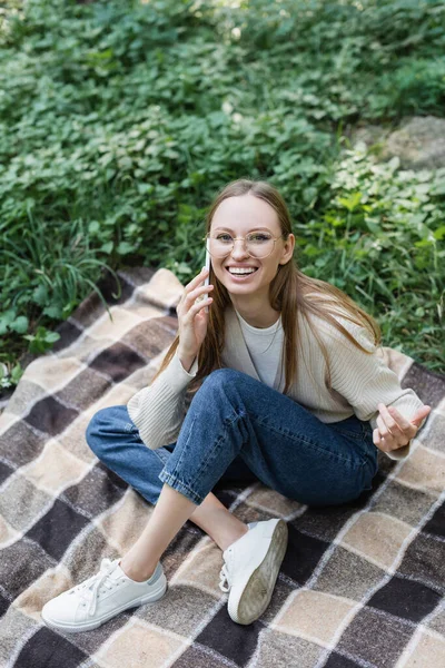 Vue grand angle de femme heureuse dans des lunettes parlant sur smartphone tout en étant assis sur carreaux plaid — Photo de stock