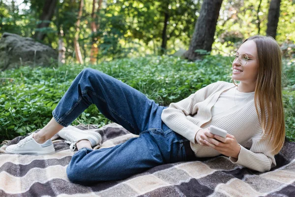 Smiling woman in glasses holding smartphone while lying on checkered plaid in green park - foto de stock