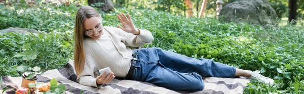 Happy woman in glasses having video call on smartphone lying on plaid blanket, banner — Stock Photo