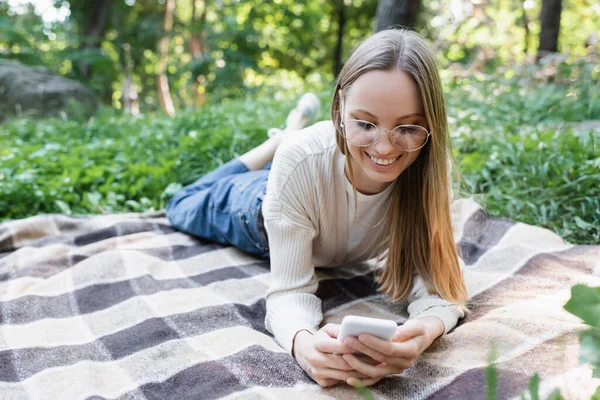 Happy woman in glasses messaging on smartphone while lying on plaid blanket in park — Stock Photo