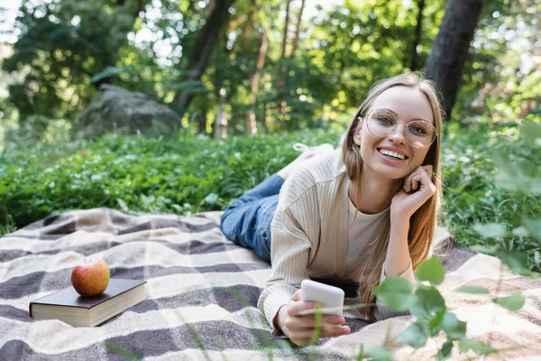 Mujer feliz en gafas sosteniendo el teléfono inteligente mientras se encuentra cerca de la manzana y libro a cuadros a cuadros - foto de stock