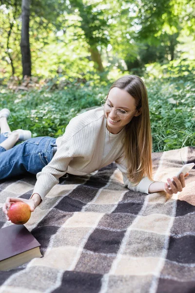 Happy woman in glasses holding smartphone while taking apple on checkered plaid — Foto stock