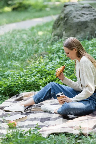 Side view of happy woman in glasses holding paper cup and croissant while sitting on blanket during picnic — Stock Photo