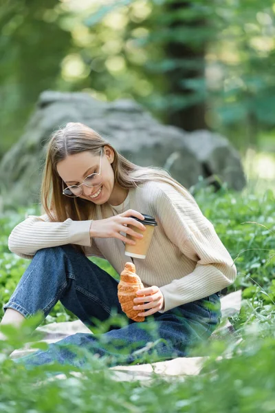 Joyful woman in glasses holding paper cup and croissant during picnic — Stock Photo