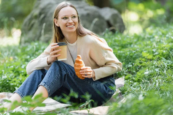 Smiling woman in glasses holding paper cup and croissant during picnic in green park — Stock Photo