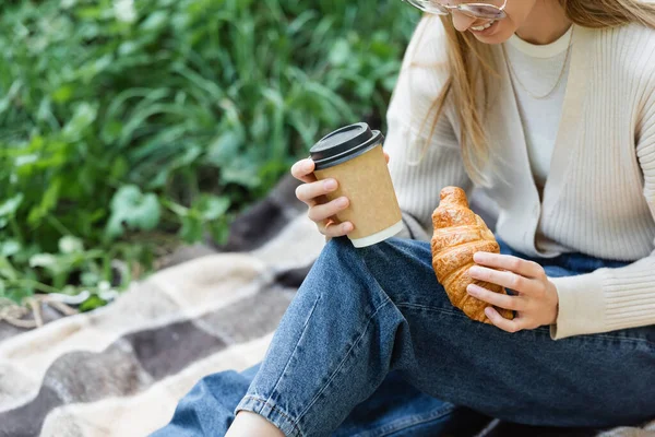 Vista recortada de la mujer feliz en gafas sosteniendo taza de papel y croissant mientras está sentado en la manta durante el picnic - foto de stock