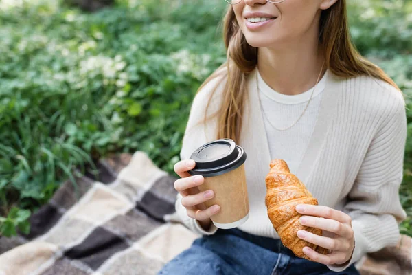 Cropped view of cheerful woman holding paper cup and croissant during picnic in park — Stock Photo