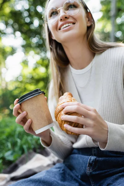 Vista basso angolo di donna felice in bicchieri che tengono tazza di carta e croissant nel parco verde — Foto stock