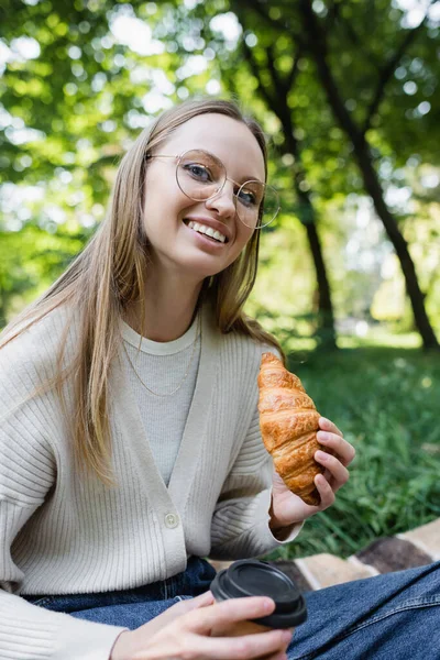 Happy woman in glasses holding tasty croissant in green park - foto de stock