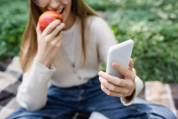 Обрезанный вид размытой женщины кусающей яблоко и делающей селфи на смартфоне — стоковое фото