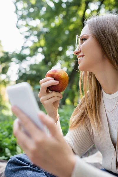 Счастливая женщина в очках с яблоком и смартфоном в руках — стоковое фото