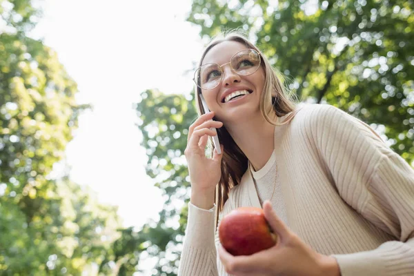 Low angle view of happy woman in glasses holding apple and talking on smartphone — Stock Photo