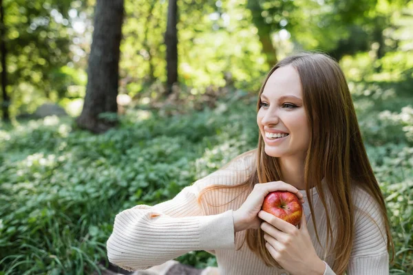 Cheerful woman holding red tasty apple in green park — стоковое фото