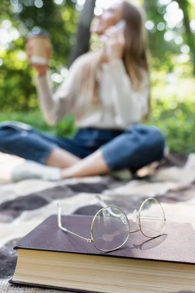 Glasses on book and plaid blanket near blurred woman during picnic — Foto stock