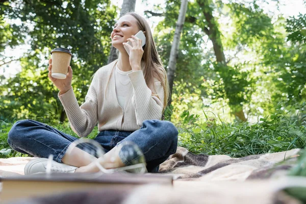 Щаслива жінка в окулярах розмовляє на смартфоні, сидячи з паперовою чашкою на картатому племені — стокове фото