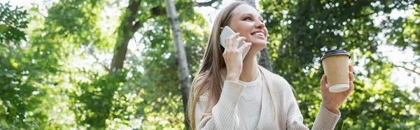 Happy woman in glasses talking on smartphone while holding paper cup in park, banner — Stock Photo