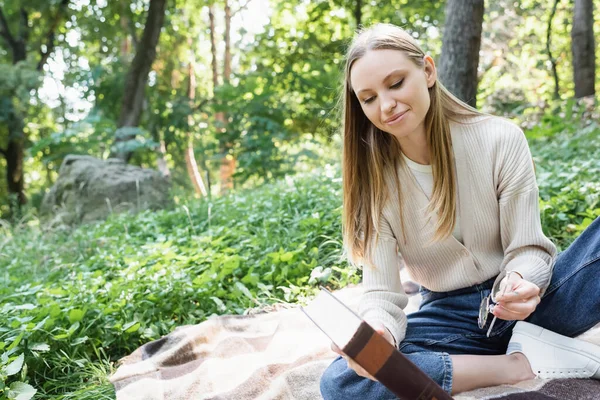 Happy woman holding book and glasses while sitting on checkered blanket — Stock Photo