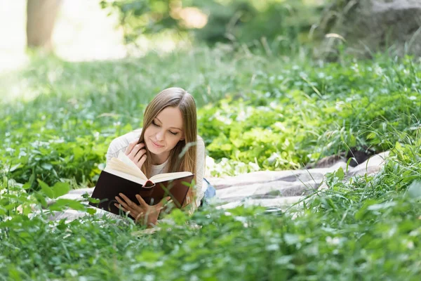 Happy woman reading book while lying on blanket around green grass — Stock Photo