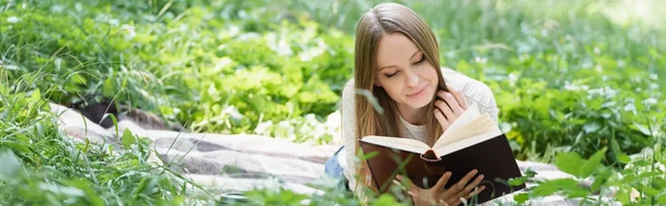 Happy woman reading book while lying on blanket around green grass, banner — Stock Photo