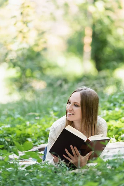Весела жінка читає книгу, лежачи на ковдрі навколо зеленої трави — стокове фото