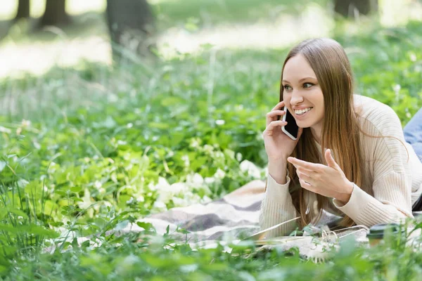 Cheerful woman talking on cellphone while lying on blanket around green grass — Stock Photo