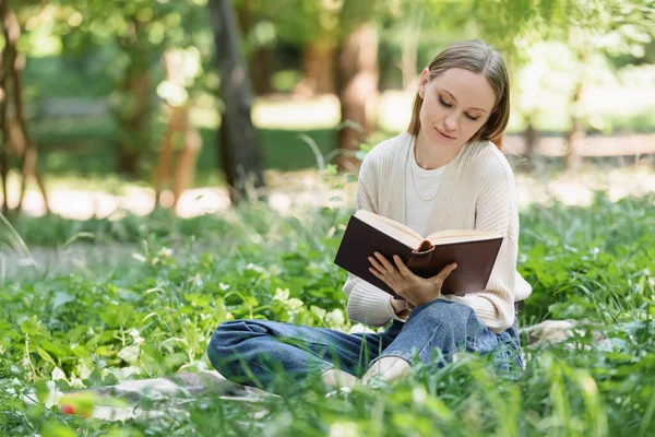 Frau liest Roman, während sie auf einer Decke im grünen Gras im Park sitzt — Stockfoto