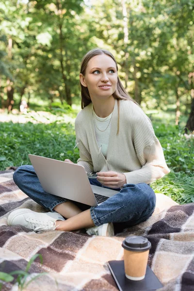 Happy freelancer sitting on blanket with crossed legs and laptop while holding glasses — Foto stock