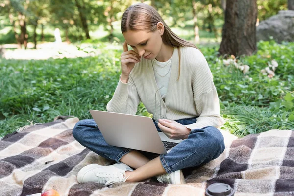 Freelancer sitting on blanket with crossed legs and using laptop while holding glasses — Photo de stock