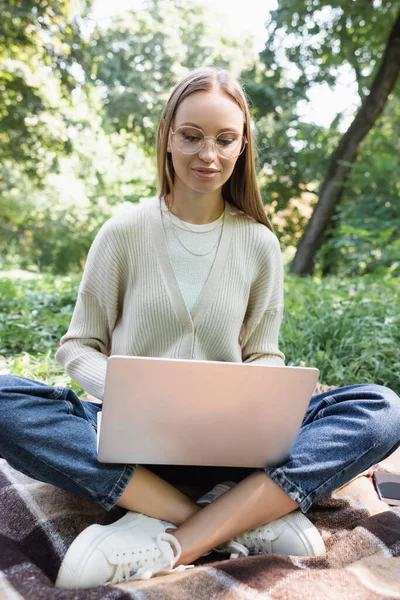 Freelancer en gafas sentado sobre manta con las piernas cruzadas y usando laptop - foto de stock