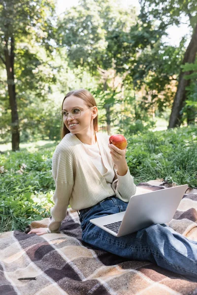 Улыбающийся фрилансер в очках, сидящий на одеяле с спелым яблоком и ноутбуком — стоковое фото