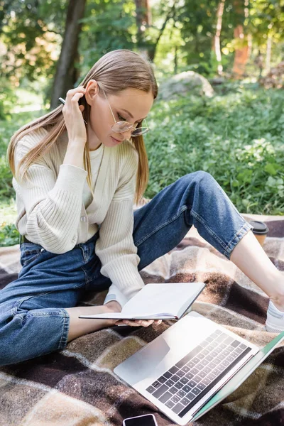 Freelancer in glasses sitting on blanket and looking at notebook near laptop — Stock Photo