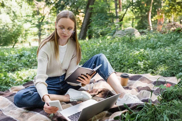 Freelancer in glasses sitting on blanket and looking at smartphone near laptop - foto de stock