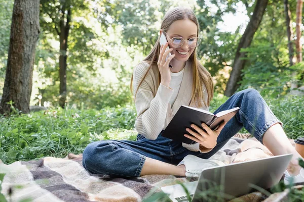 Happy freelancer in glasses holding notebook while sitting on blanket and talking on smartphone near laptop — Foto stock