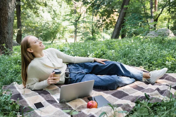 Heureux pigiste dans des lunettes tenant tasse en papier tout en se reposant sur la couverture près de gadgets — Photo de stock