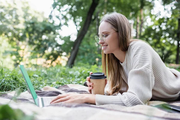 Happy freelancer in glasses holding paper cup while lying on blanket and using laptop in park — Stockfoto