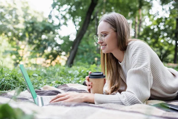 Feliz freelancer en gafas sosteniendo la taza de papel mientras está acostado en la manta y el uso de ordenador portátil en el parque - foto de stock