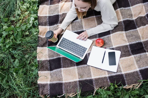 Вид сверху на фрилансера в очках лежащего на одеяле и использующего ноутбук в парке — стоковое фото