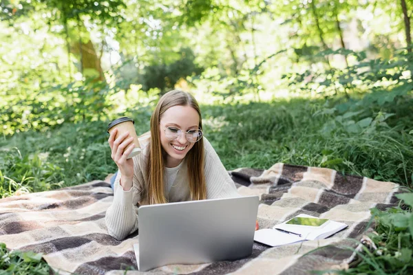 Femme souriante dans des lunettes allongées sur une couverture, tenant une tasse en papier et utilisant un ordinateur portable dans le parc — Photo de stock