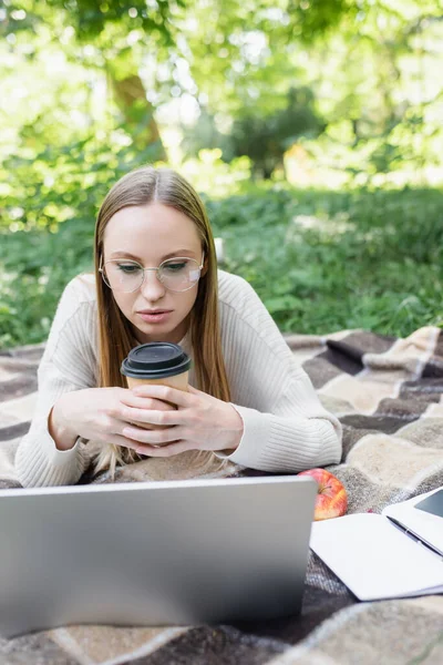 Frau in Brille liegt auf Decke, hält Pappbecher in der Hand und benutzt Laptop im Park — Stockfoto