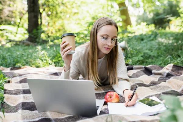 Femme dans des lunettes prendre des notes tout en tenant tasse de papier près de gadgets sur couverture dans le parc — Photo de stock