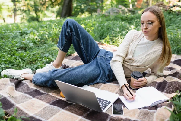 Donna in occhiali che prende appunti mentre tiene la tazza di carta vicino al laptop e allo smartphone sulla coperta nel parco — Foto stock