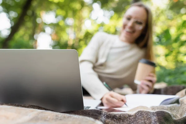 Laptop on blanket near blurred woman with paper cup in park — Foto stock