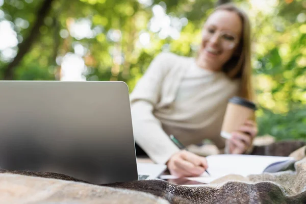 Laptop on blanket near blurred woman with paper cup in park — Stock Photo