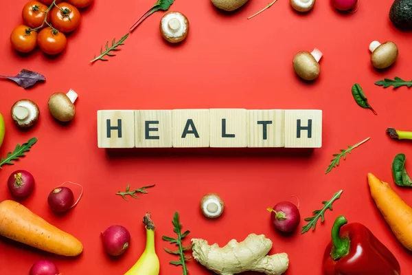 Poser à plat avec des lettres de santé sur des cubes de bois près de légumes et fruits biologiques sur fond rouge — Photo de stock