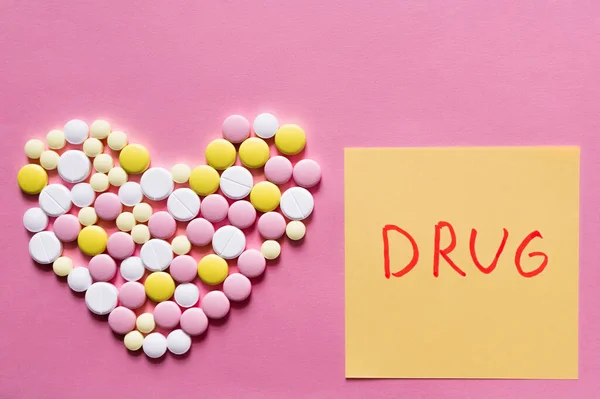 Piatto lay vista di diverse pillole rotonde a forma di cuore vicino nota di carta con lettering droga su sfondo rosa — Foto stock