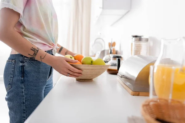 Vista ritagliata della donna tatuata che tiene la ciotola con le mele vicino alla scatola del pane e alla caffettiera offuscata — Foto stock