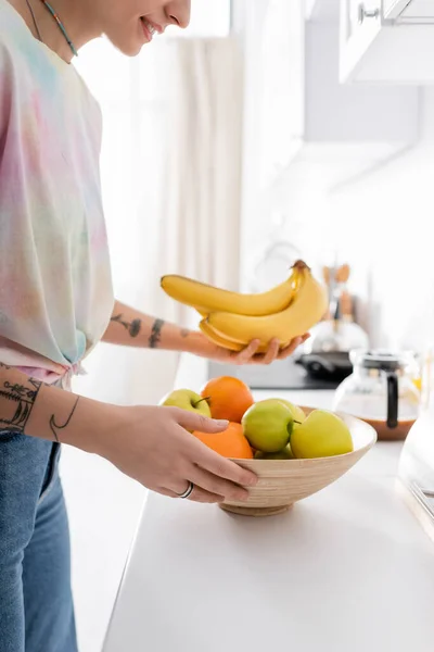 Частковий вид татуйованої жінки, що тримає стиглі банани біля миски з яблуками на кухні — стокове фото